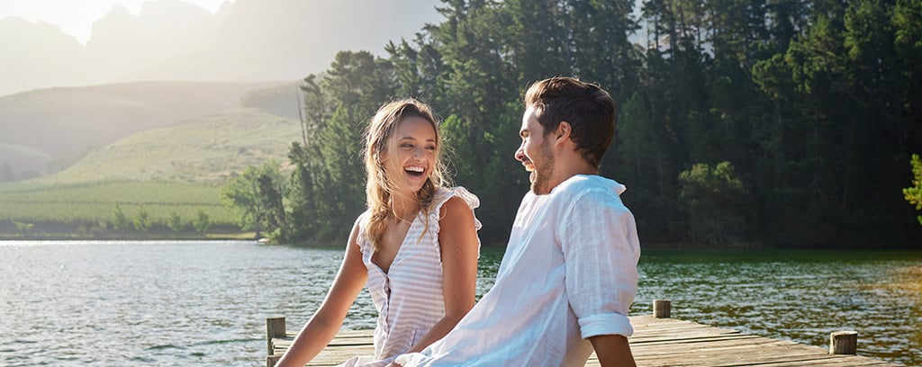 Love, laugh and pier with couple at lake for bonding, romance and affectionate date. Nature, travel and holiday with man and woman sitting on boardwalk in countryside for happy, summer and vacation